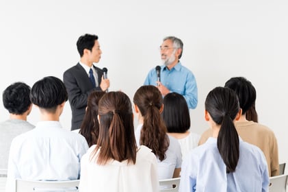 講演会のステージ上でマイクを持ち、落ち着いた表情で司会進行を行う担当者の様子